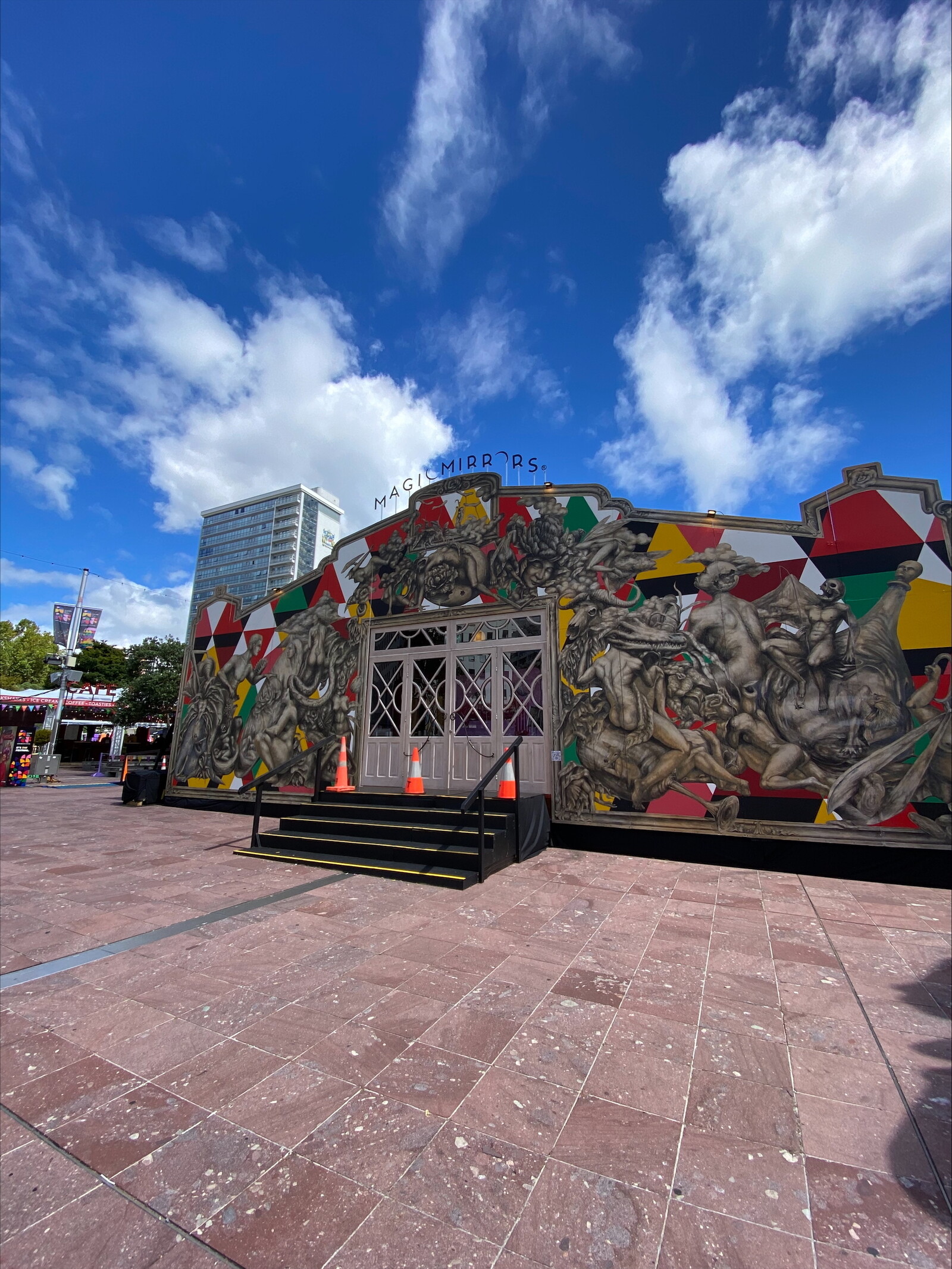 A vibrant Spiegeltent stands against a bright blue sky with scattered clouds. The ornate, temporary structure features a striking mural with intricate black-and-white illustrations of human figures intertwined, accented by geometric patterns in red, yellow, and green. The entrance has elegant double doors framed by glass panels, with black steps leading up to it, marked by safety cones. Above the doorway, a sign reads "MAGIC MIRRORS," adding a whimsical touch. In the background, a tall modern building contrasts with the historical aesthetic of the Spiegeltent, while event stalls and banners hint at a lively festival atmosphere.