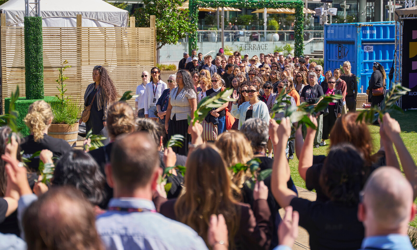 A crowd gathers outdoors for a pōwhiri, with people holding leafy branches in welcome.