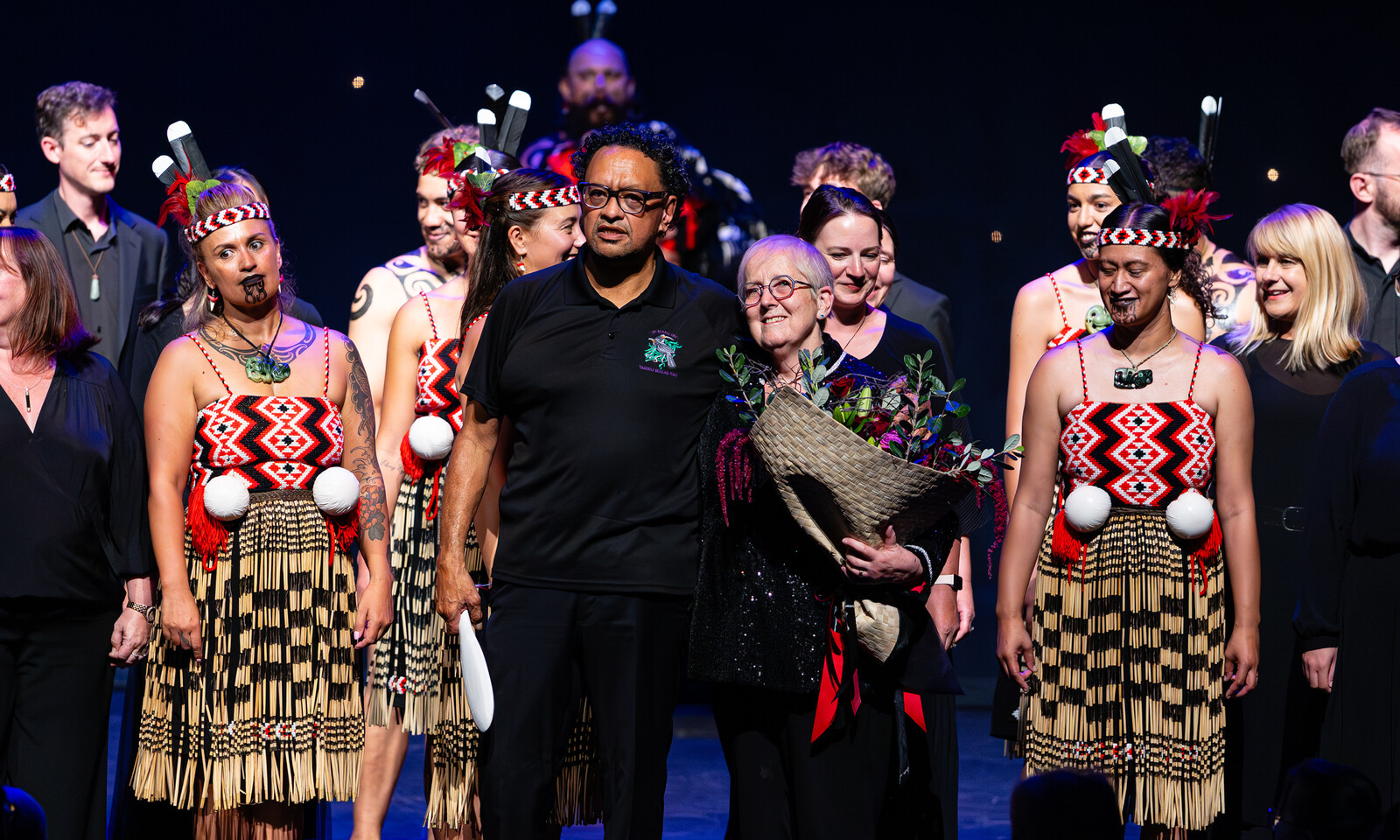 Performers and choir members stand together on stage following a performance, with flowers presented in celebration.