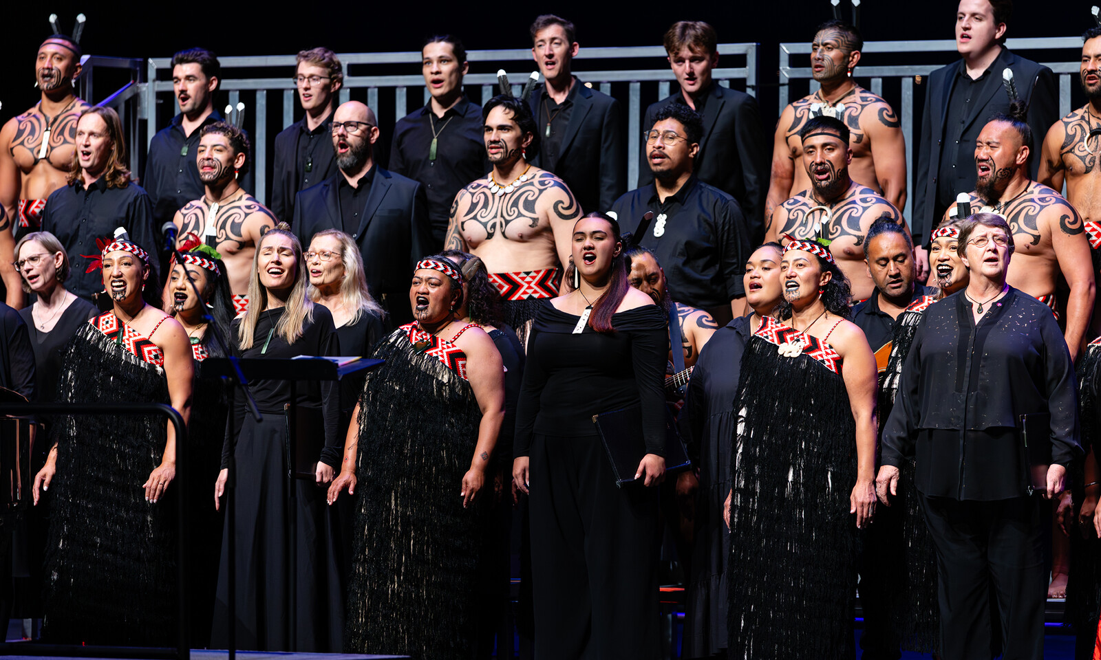 A choir performs on stage in traditional Māori attire and contemporary dress.