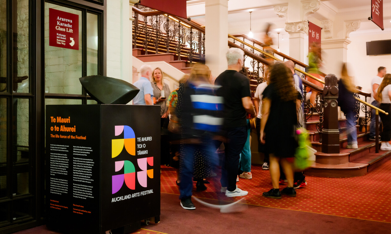 People gather in a theatre foyer beside Auckland Arts Festival signage and a grand staircase.